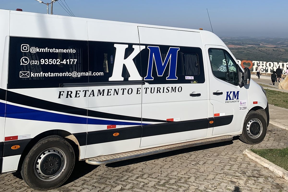 Van da KM Fretamento e Turismo estacionada em um mirante, com céu azul e o letreiro de São Thomé das Letras ao fundo